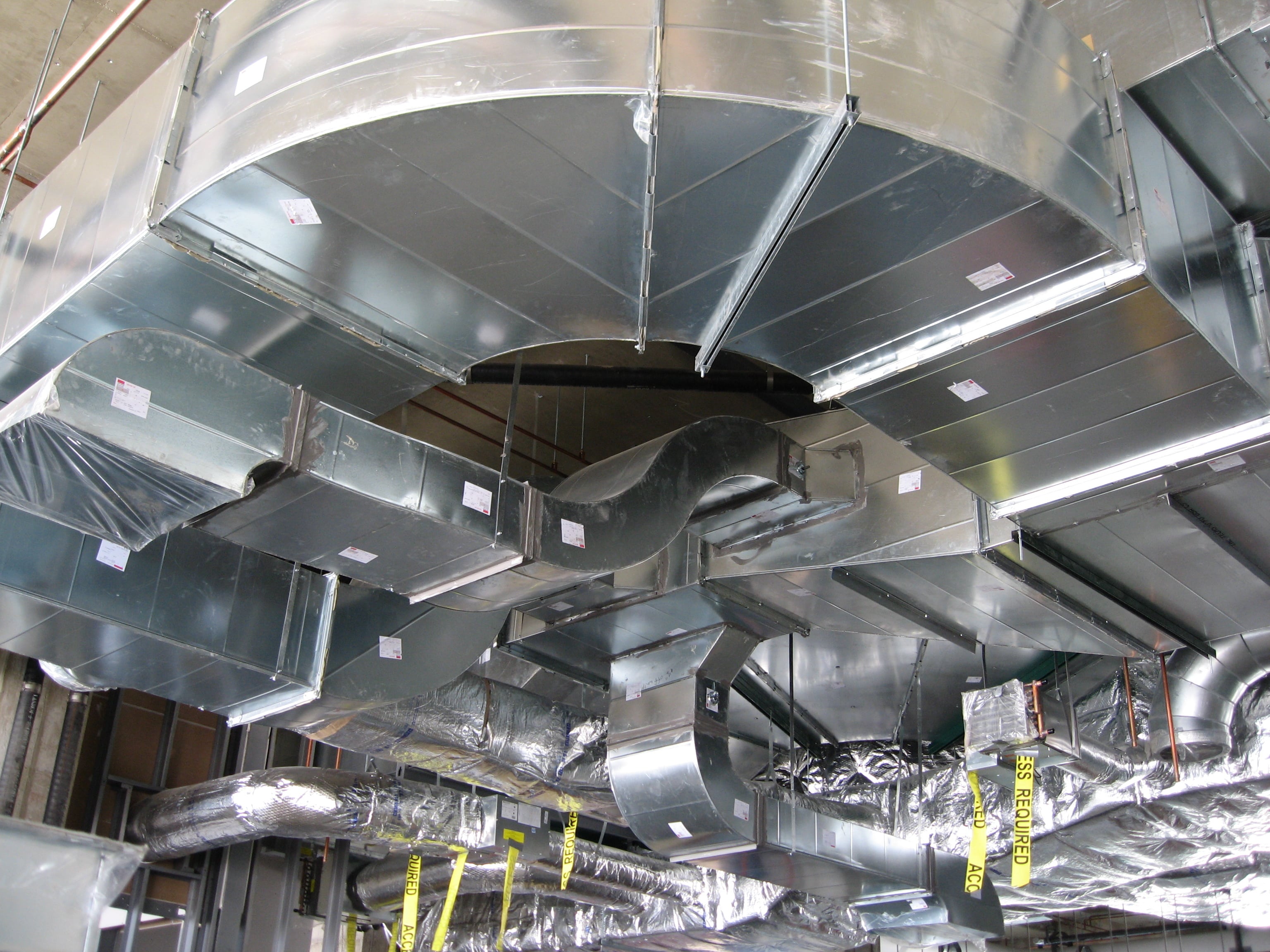 A complex arrangement of metal air ducts and tubing suspended from a ceiling, with printed labels attached for identification.