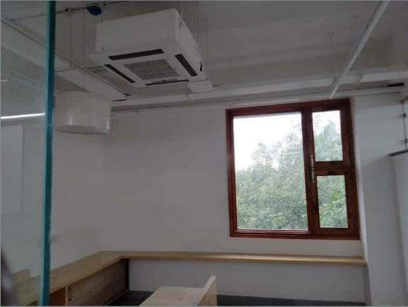 A minimalist office space with a wooden bench, large window, ceiling air conditioner, and natural light filtering in from outside.