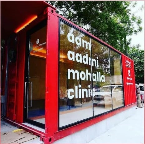 Exterior view of a vibrant red Aam Aadmi Mohalla Clinic, featuring large, white lettering on glass windows amidst greenery.