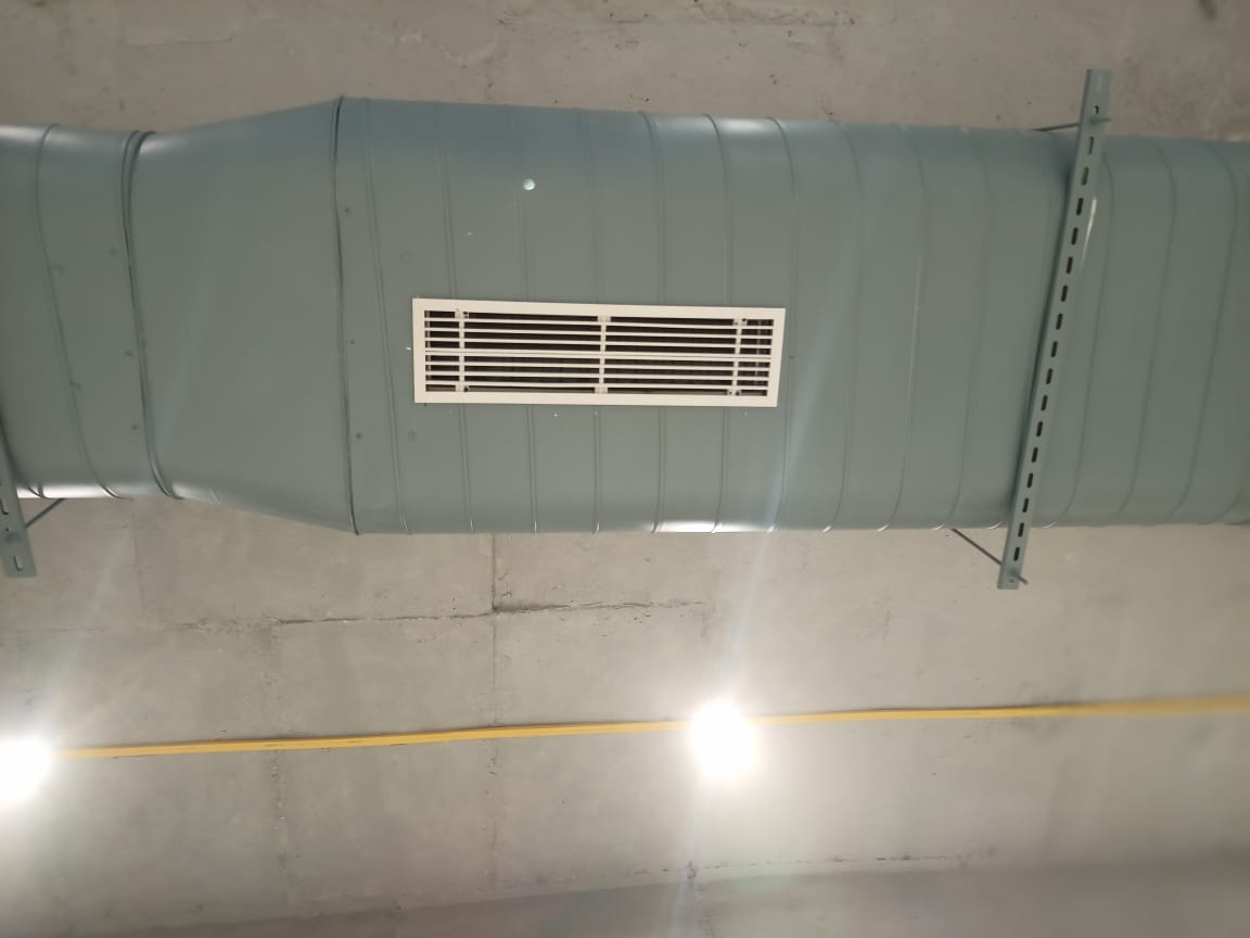 Close-up of a blue metal ventilation duct with a white vent, mounted on a concrete ceiling, illuminated by overhead lights.