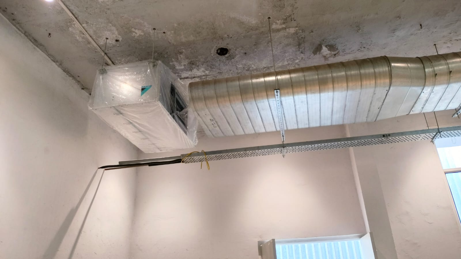 Ceiling with air conditioning unit wrapped in plastic and large metal ductwork in an unfinished room with white walls.
