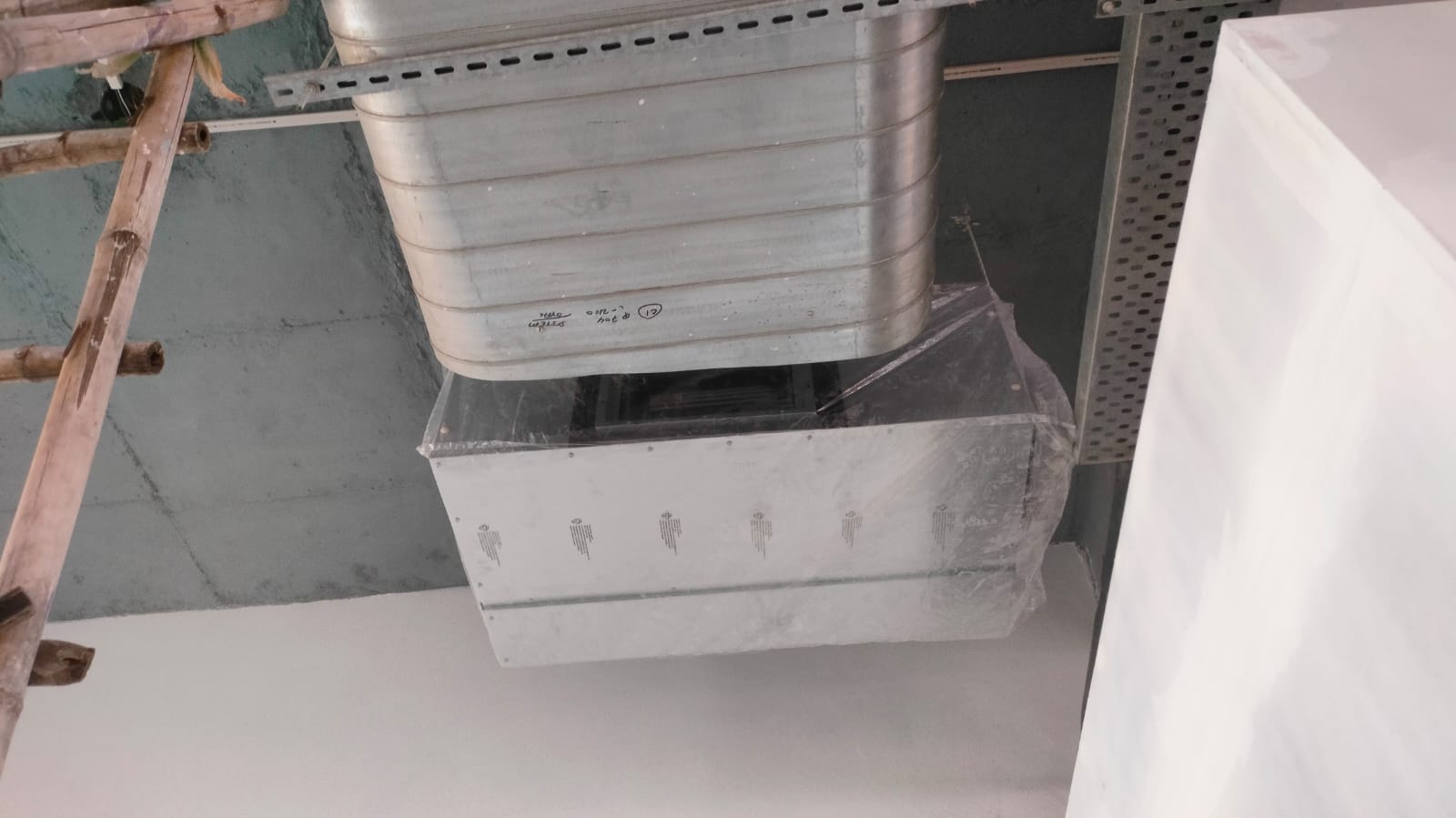 Ceiling-mounted ductwork with a plastic cover, supported by wooden scaffolding, against a partially painted wall.
