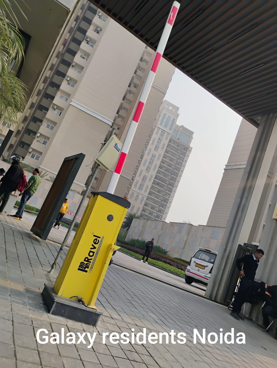 A gated entrance at Galaxy residential complex in Noida, featuring a yellow entry system, security personnel, and modern buildings in the background.