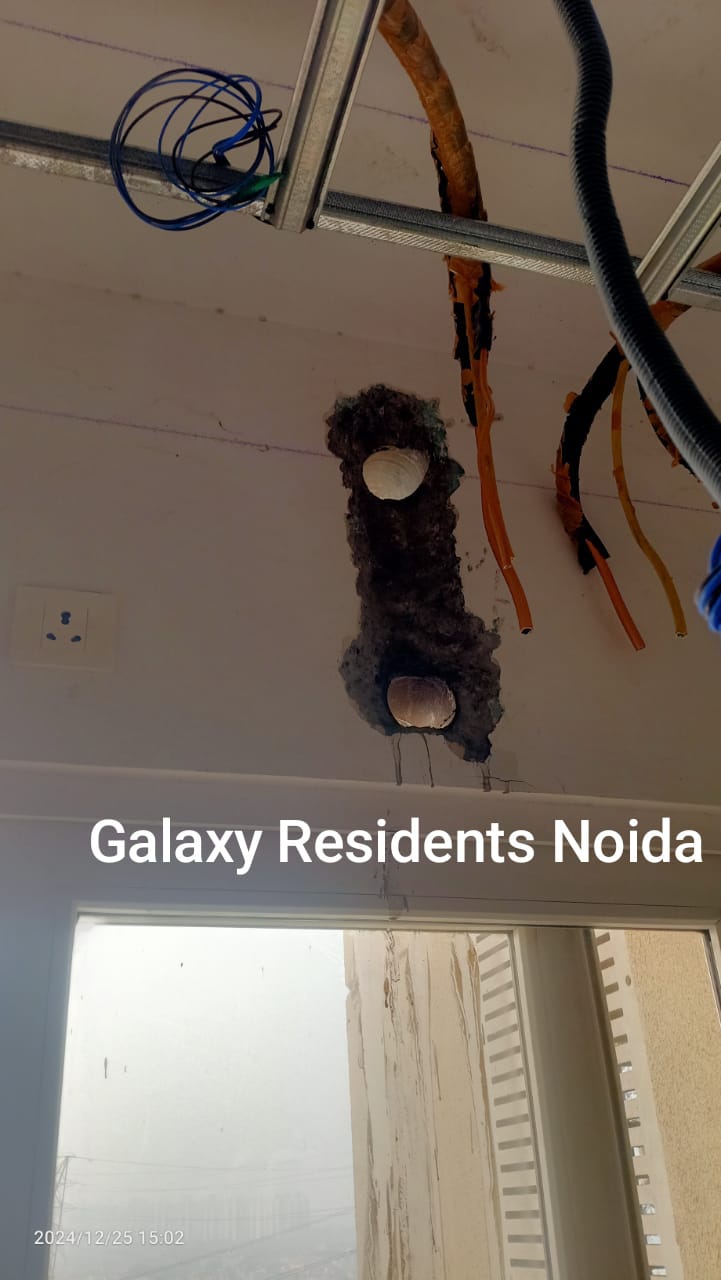 Exposed ceiling with electrical wires and two light fixture holes. Background shows a dirty window and foggy view, labeled Galaxy Residents Noida.