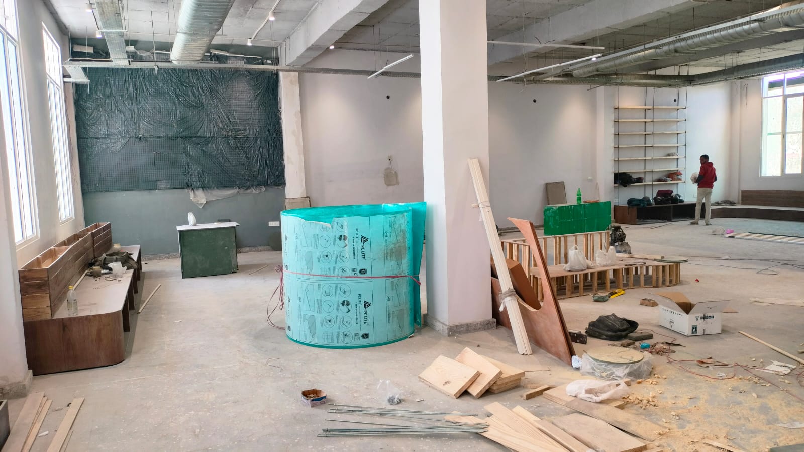 A spacious, unfinished room with construction materials, wooden benches, and a worker in red, preparing for final touches.
