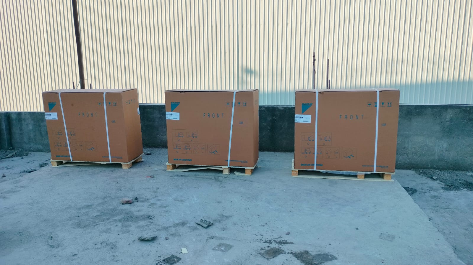 Three large, sealed cardboard boxes labeled FRONT, resting on wooden pallets against a textured concrete wall.
