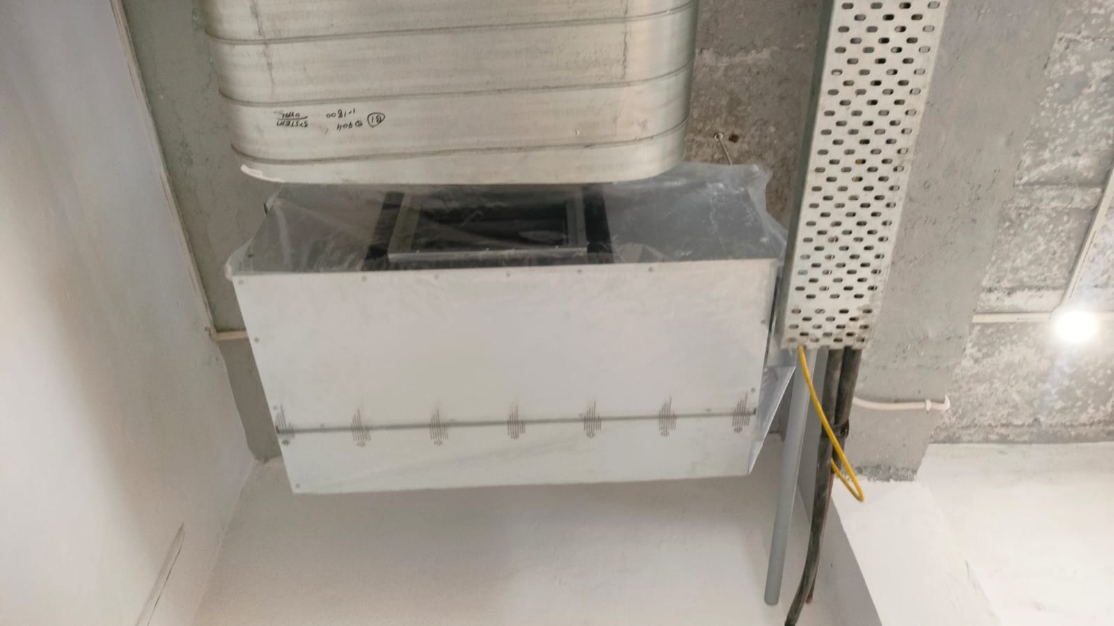 Ceiling-mounted ventilation unit with a metal duct above and a rectangular enclosure below, partially covered in plastic film.